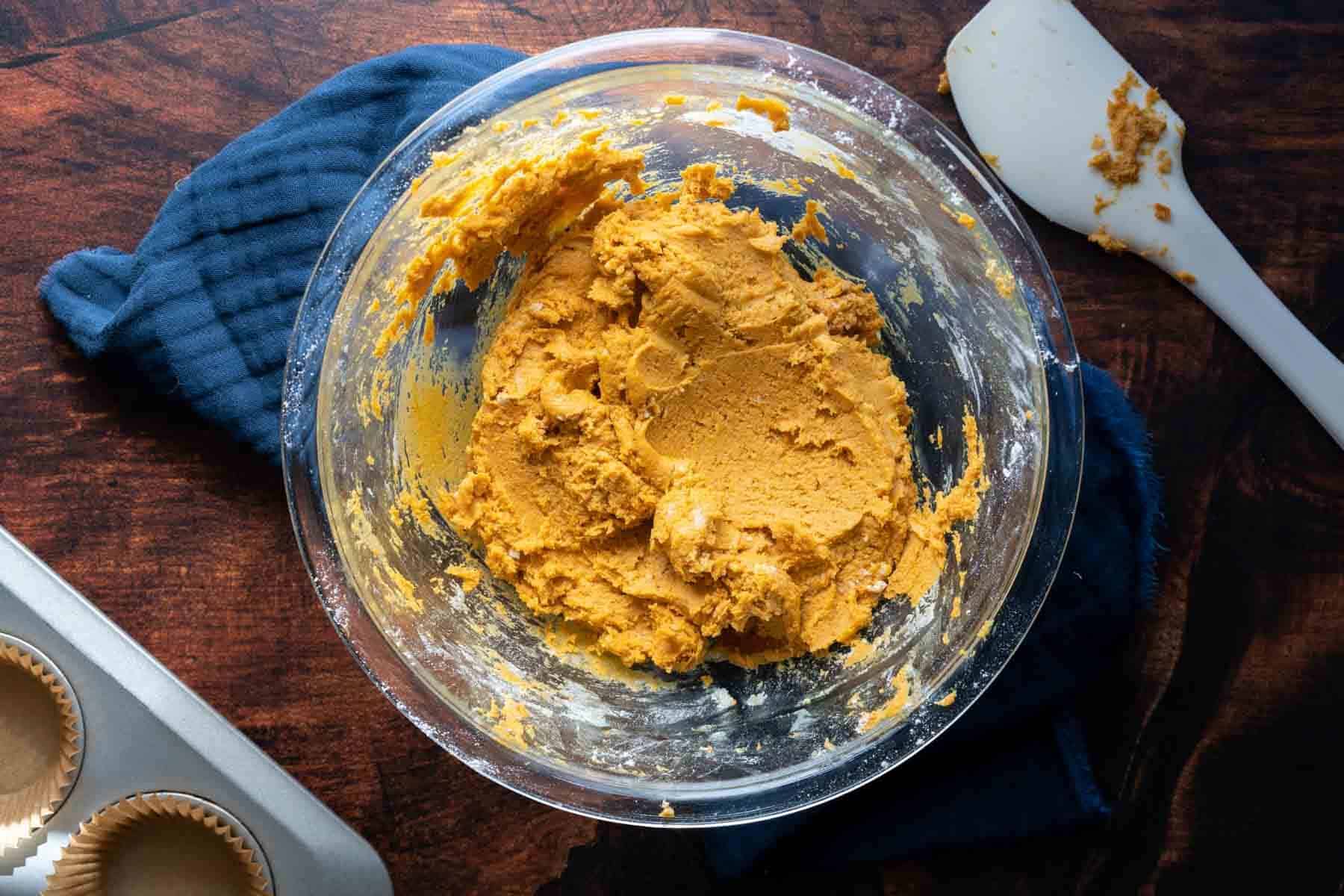 A glass bowl contains thick peanut butter and pumpkin pupcake batter, placed on a dark cloth with a spatula and a muffin tin lined with paper cups nearby on a wooden surface.