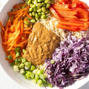 Bowl with gluten-free peanut sauce, shredded carrots, red bell pepper slices, edamame, shredded purple cabbage, sliced green onions over noodles.