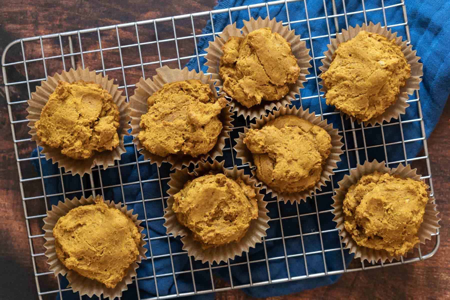 Eight pumpkin pupcakes in brown paper liners are cooling on a wire rack over a blue cloth on a wooden surface.