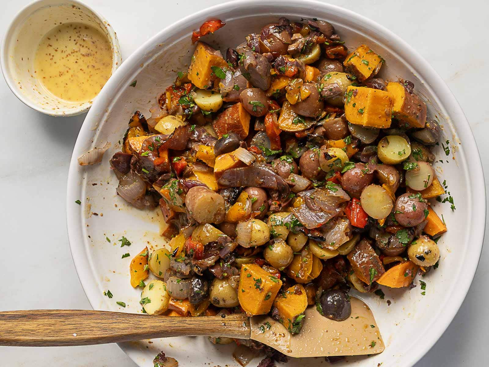 A bowl of roasted mixed vegetables, including potatoes and sweet potatoes, with herbs, offers a Rainbow Potato Salad vibe-served next to a small cup of dressing and a wooden spatula.
