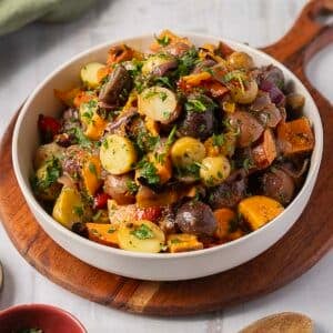 A bowl of Rainbow Potato Salad featuring mixed rainbow potatoes sweet potatoes, and vegetables, garnished with chopped parsley and placed on a wooden serving board.
