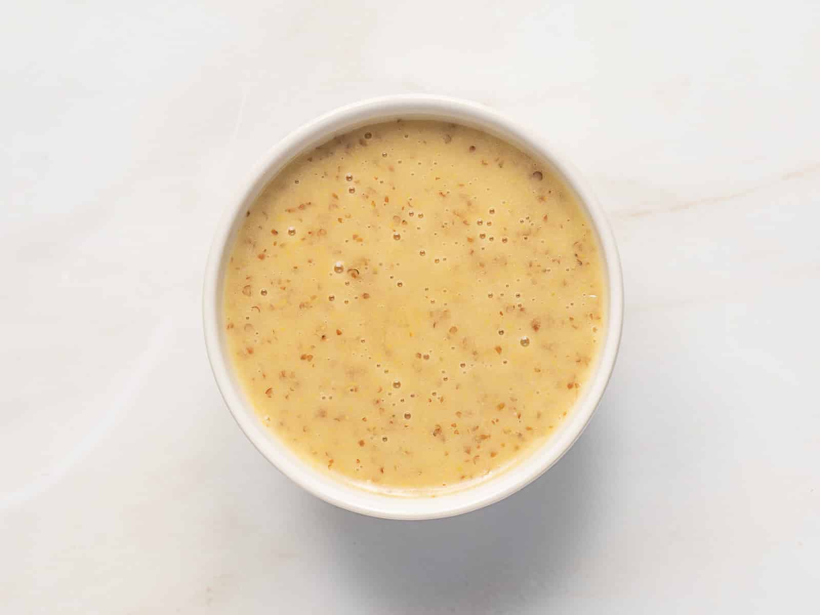 A small white bowl filled with whole grain mustard sauce, perfect for drizzling over Rainbow Potato Salad, photographed from above on a light-colored surface.