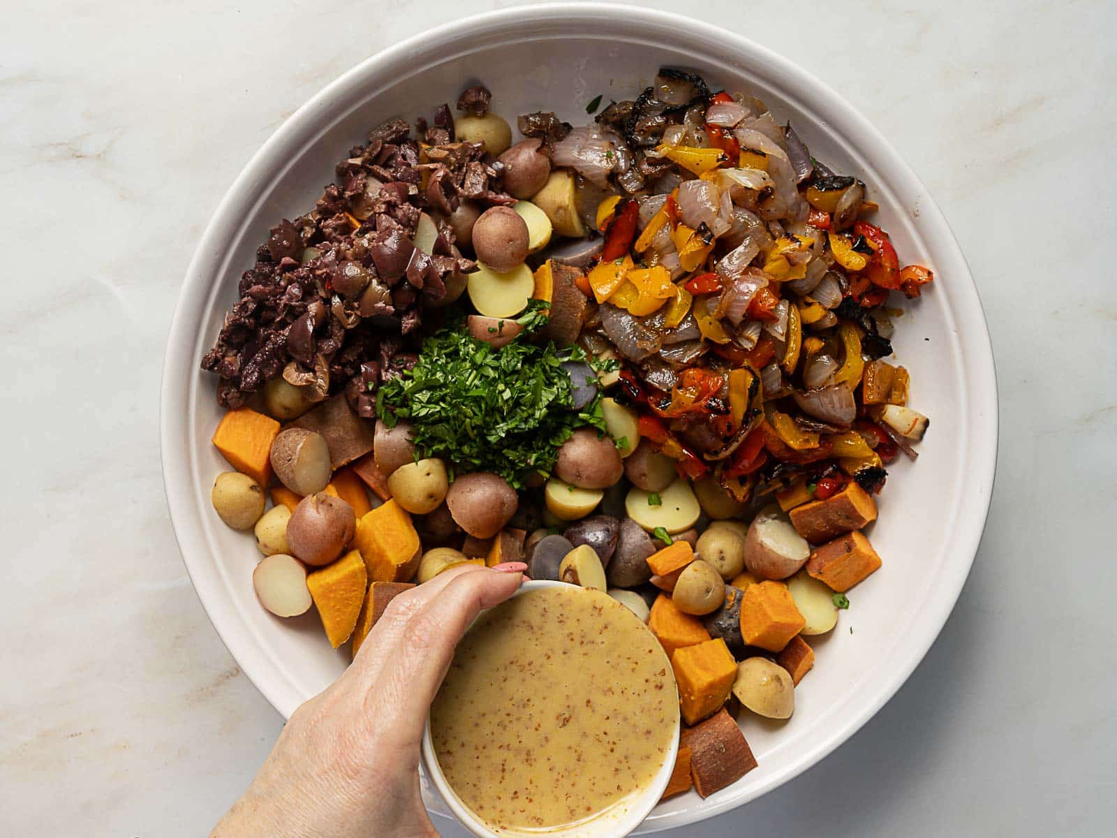 A bowl with chopped vegetables, herbs, and olives is ready for a burst of flavor as a hand pours mustard dressing over this vibrant Rainbow Potato Salad.
