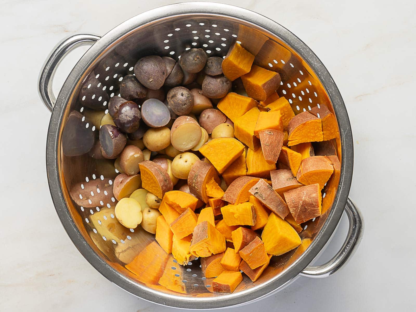 A metal colander containing halved small purple and yellow potatoes and chunks of sweet potato on a white surface, perfect for making Rainbow Potato Salad.