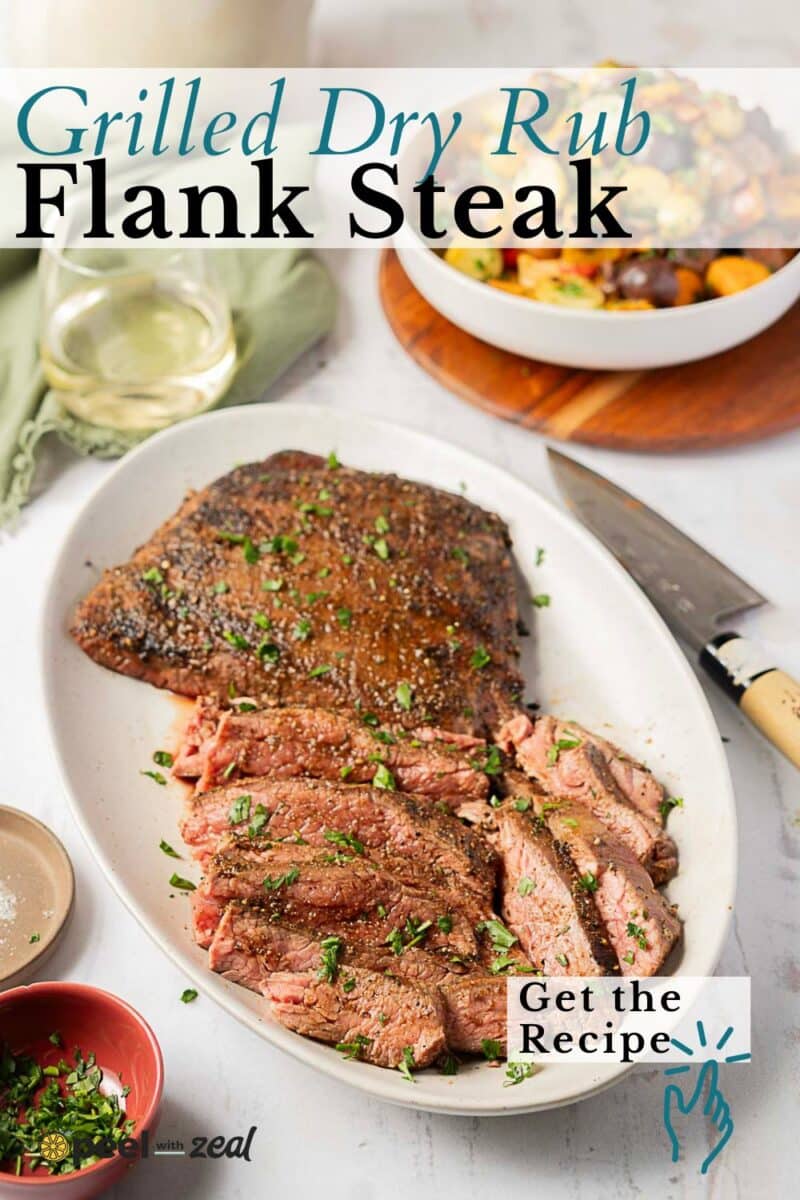 A platter of spice crusted grilled flank steak, partially sliced and garnished with herbs, sits alongside a knife, a glass of white wine, and a bowl of roasted vegetables.