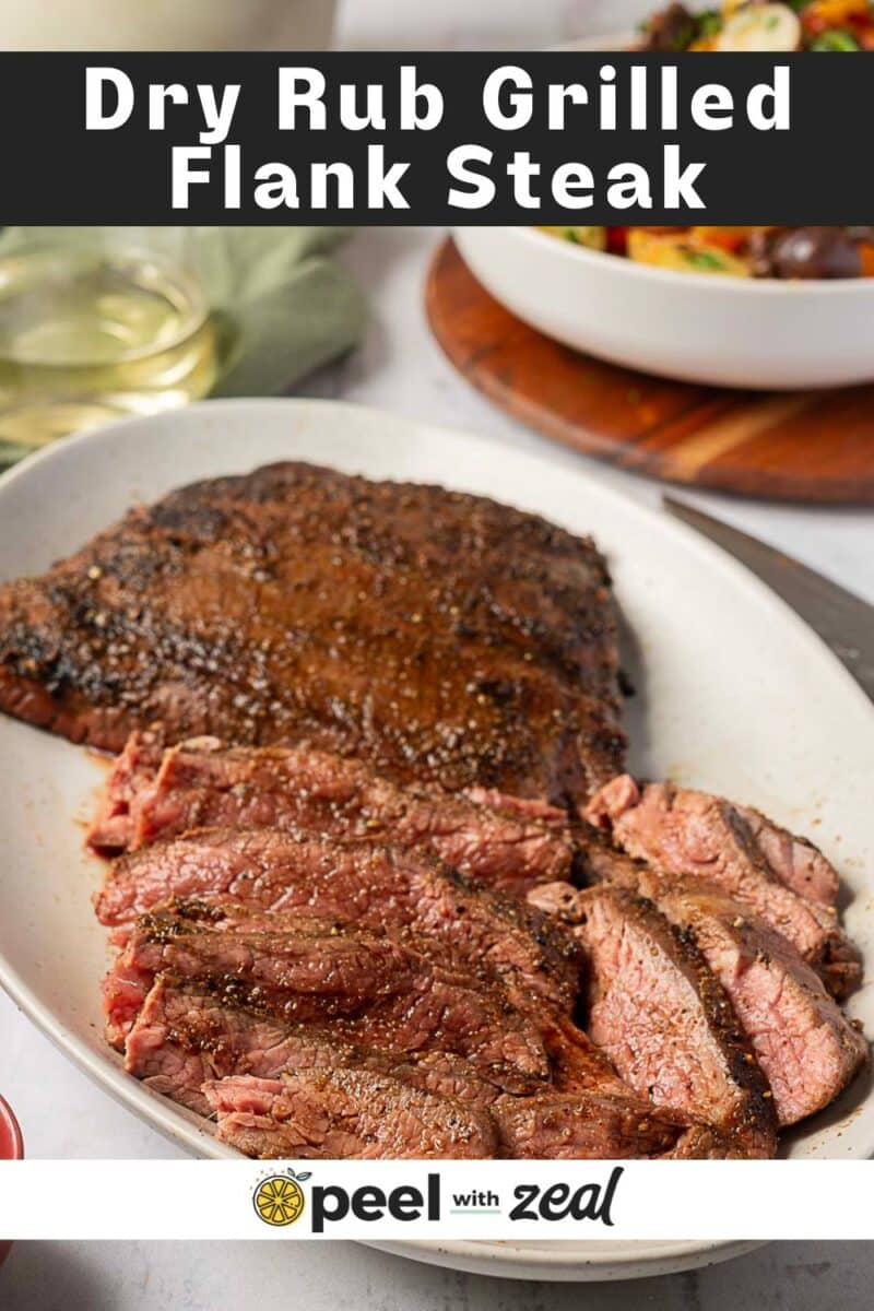 A spice crusted grilled flank steak, partly sliced, rests on an oval white plate. In the background, a bowl of salad and a glass of white wine add to the inviting scene.