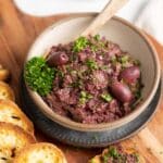 A bowl of kalamata tapenade garnished with parsley and olives sits on a wooden board, accompanied by toasted bread slices.