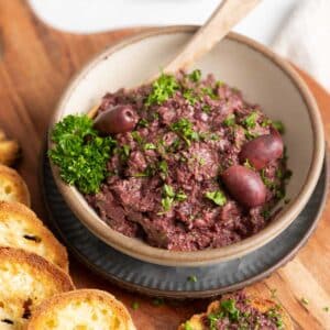 A bowl of kalamata tapenade garnished with parsley and olives sits on a wooden board, accompanied by toasted bread slices.