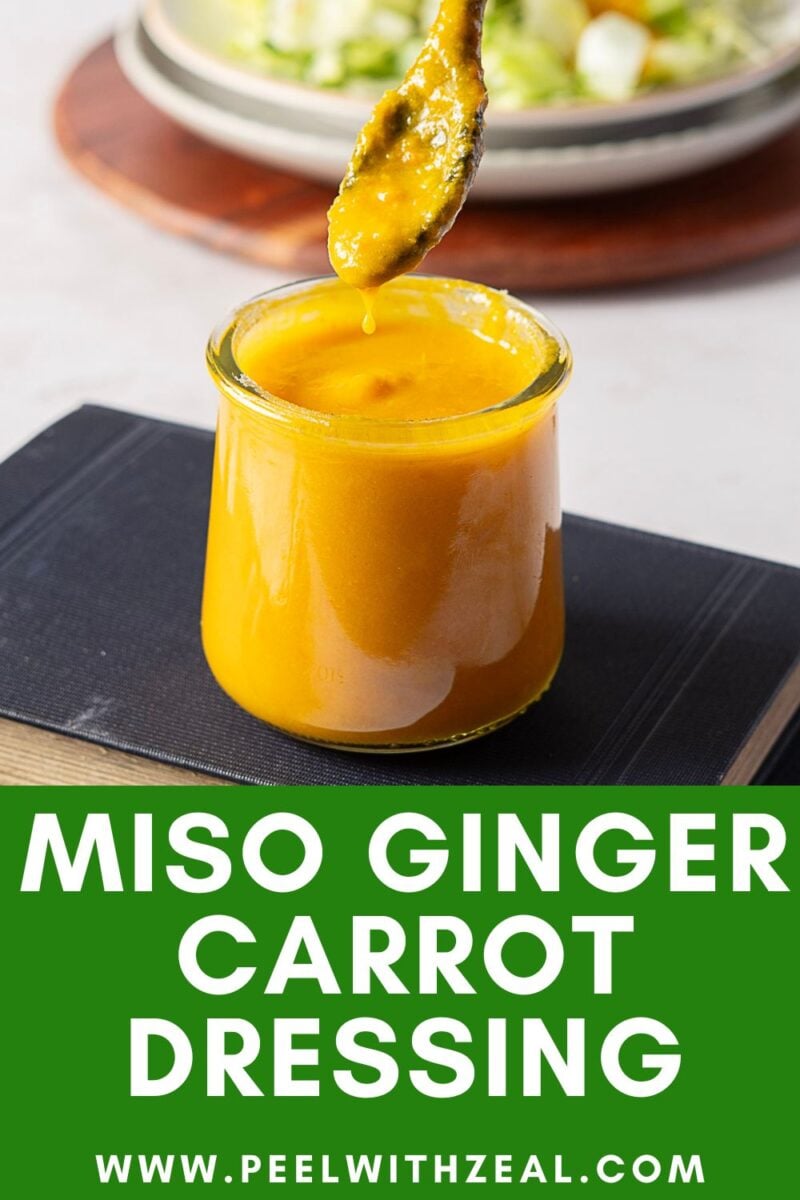 A glass jar filled with vibrant carrot ginger dressing sits atop a closed book, while a spoon drips some above the jar. Text at the bottom reads, “Miso Ginger Carrot Dressing www.peelwithzeal.com.”.