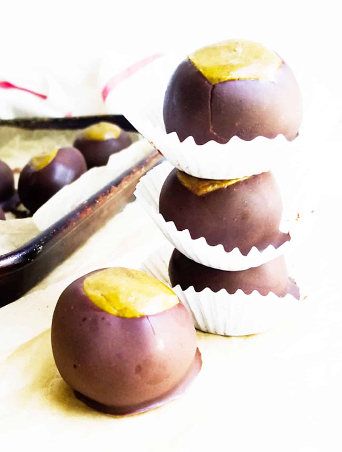 Stack of three chocolate-covered protein buckeyes in paper cups, with more on a tray in the background.