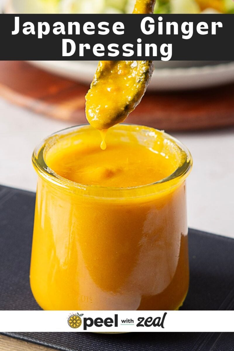 A glass jar filled with bright yellow carrot ginger dressing sits on a dark surface, with a spoon above the jar letting the dressing drip back in. The text "Japanese Ginger Dressing" is at the top.
