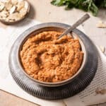 A bowl of thick red romesco sauce with a metal spoon rests on a dark circular mat. Nearby, almond slices and green leaves adorn the textured surface, scattered with paper pages.