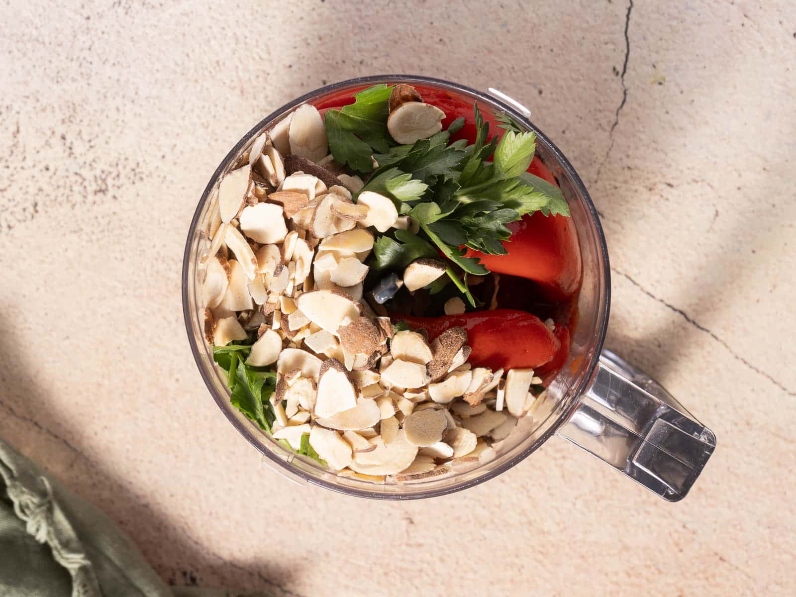 Overhead view of a blender cup containing almonds, tomatoes, parsley, and leafy greens on a textured surface, perfect for crafting a no-cook romesco sauce.