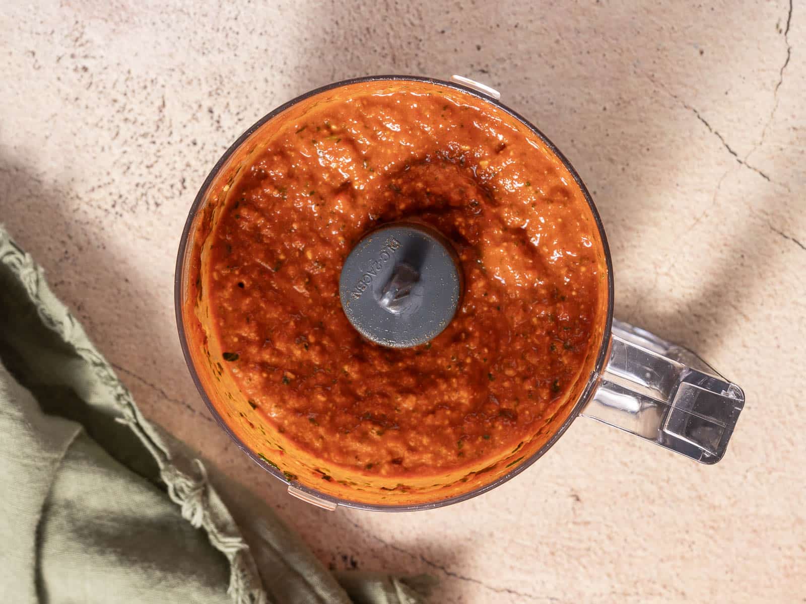 A food processor filled with no-cook romesco sauce sits on a beige surface, accompanied by a nearby green cloth.