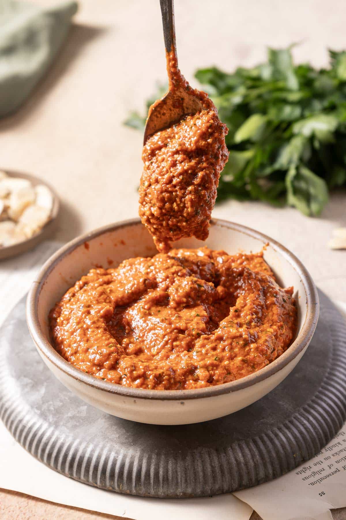 A spoon dips into a textured, romesco sauce nestled in a bowl on a plate. Fresh herbs and almonds subtly grace the background.