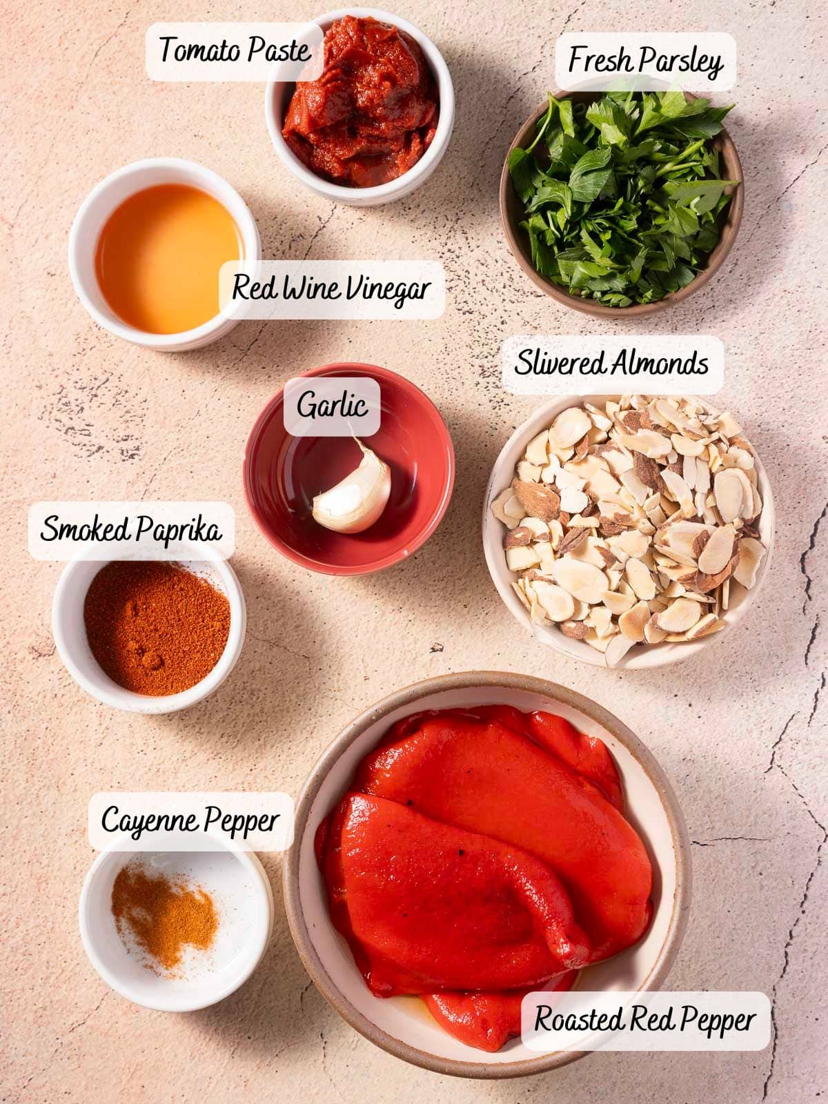 On the table are ingredients for the romesco sauce recipe: tomato paste, fresh parsley, red wine vinegar, slivered almonds, garlic, smoked paprika, cayenne pepper, and roasted red pepper.