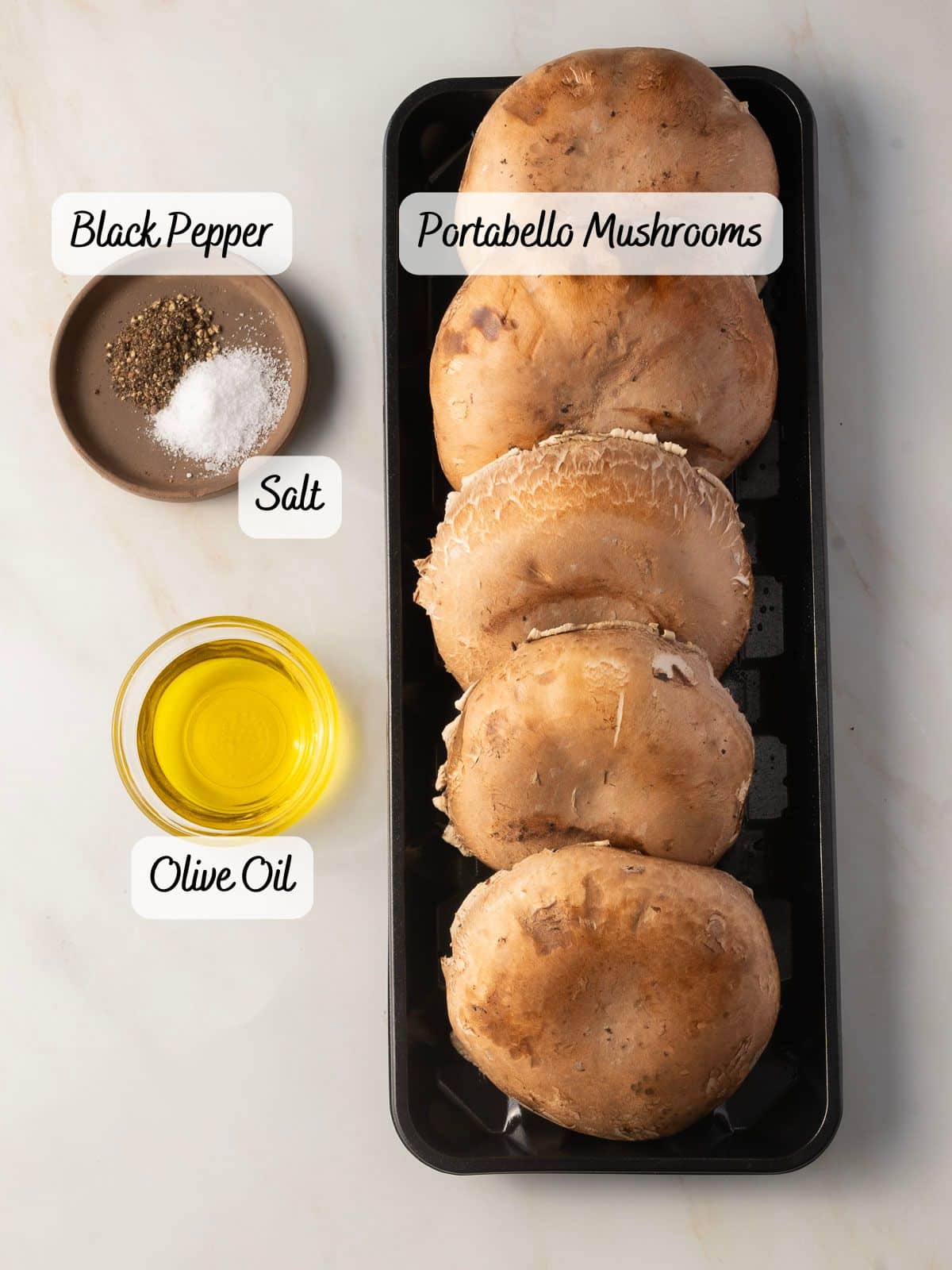 Overhead view of five roasted portabella mushrooms in a tray with labeled ingredients: black pepper, salt, and olive oil on the side.
