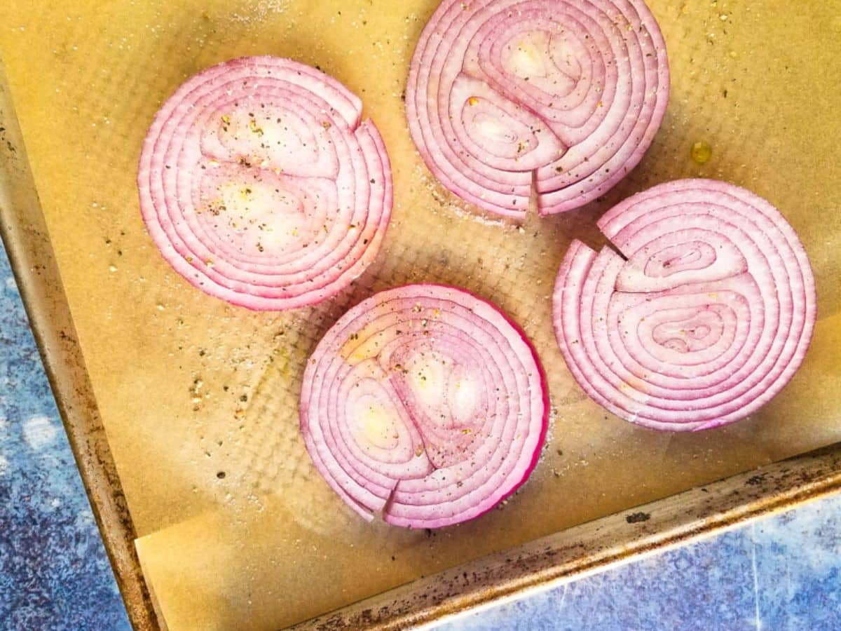 Four sliced red onions, perfectly seasoned with pepper and ready to be roasted, sit on parchment paper atop a baking tray.