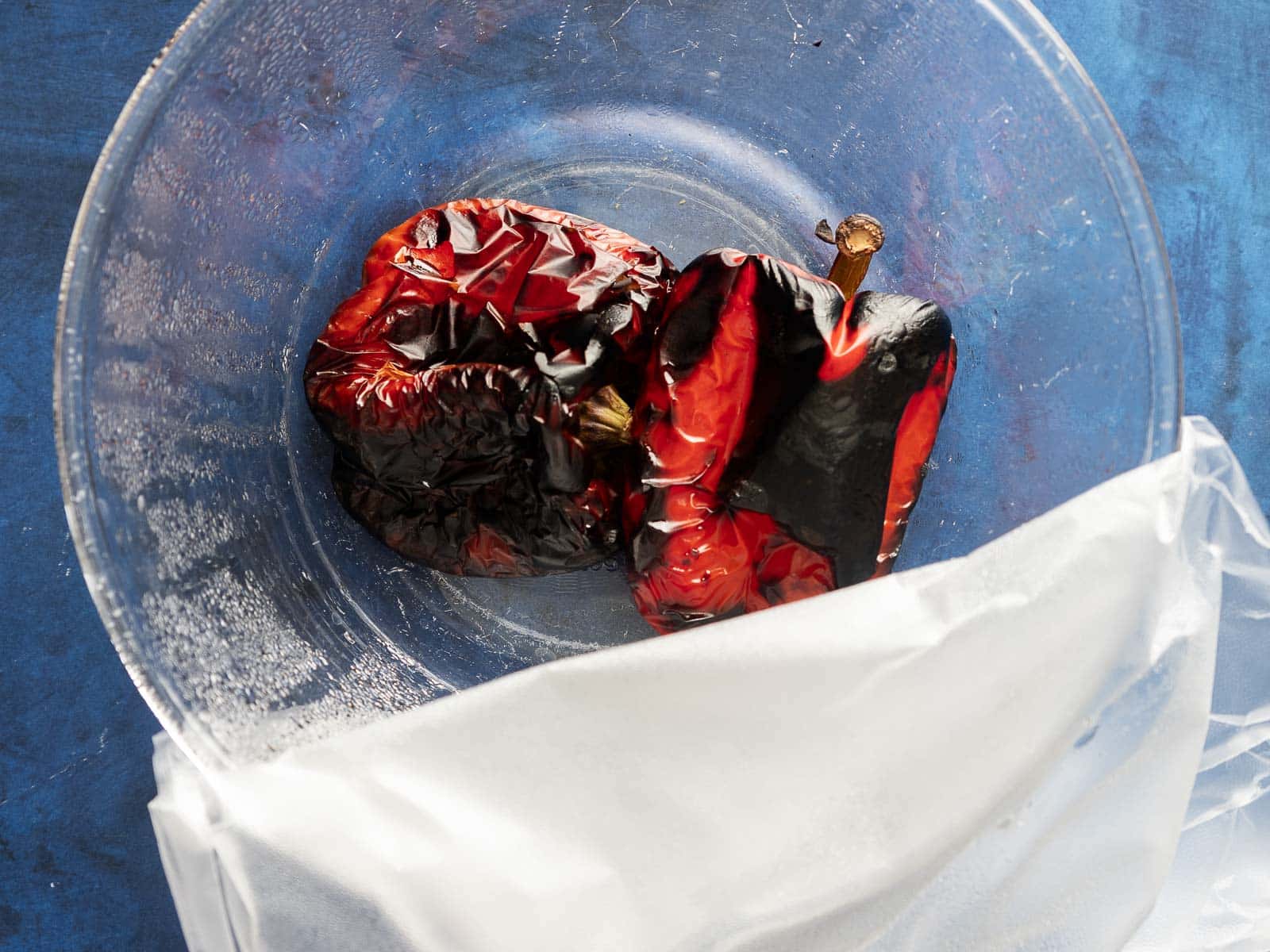 Roasted red peppers in a clear bowl with a white plastic bag on a blue surface.
