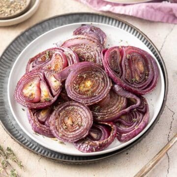 A beautifully arranged plate of roasted red onions, perfectly seasoned with herbs, sits invitingly on a white and black rimmed plate.