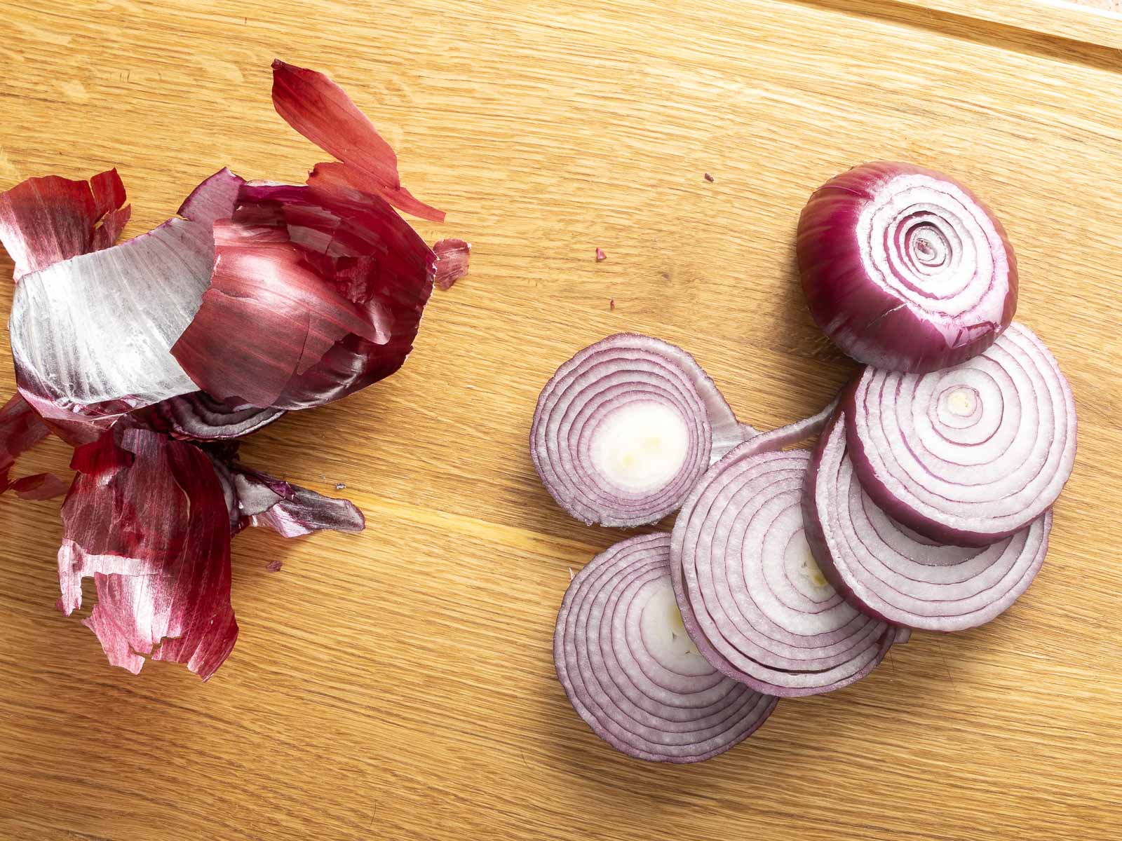 Sliced red onion and onion skin on a wooden cutting board, ready to become delicious roasted red onions.