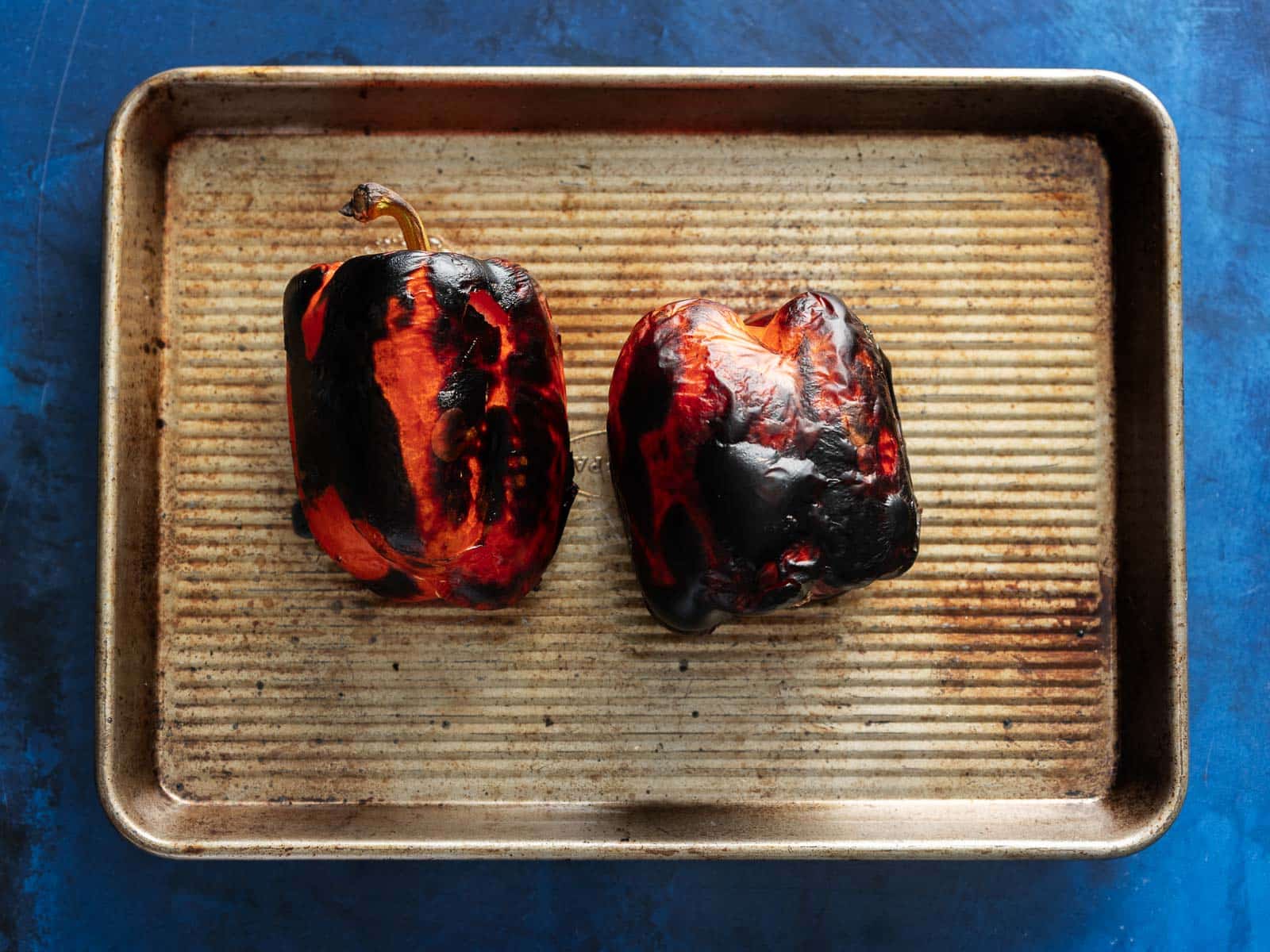 Two roasted red bell peppers rest on a baking sheet against a blue background.