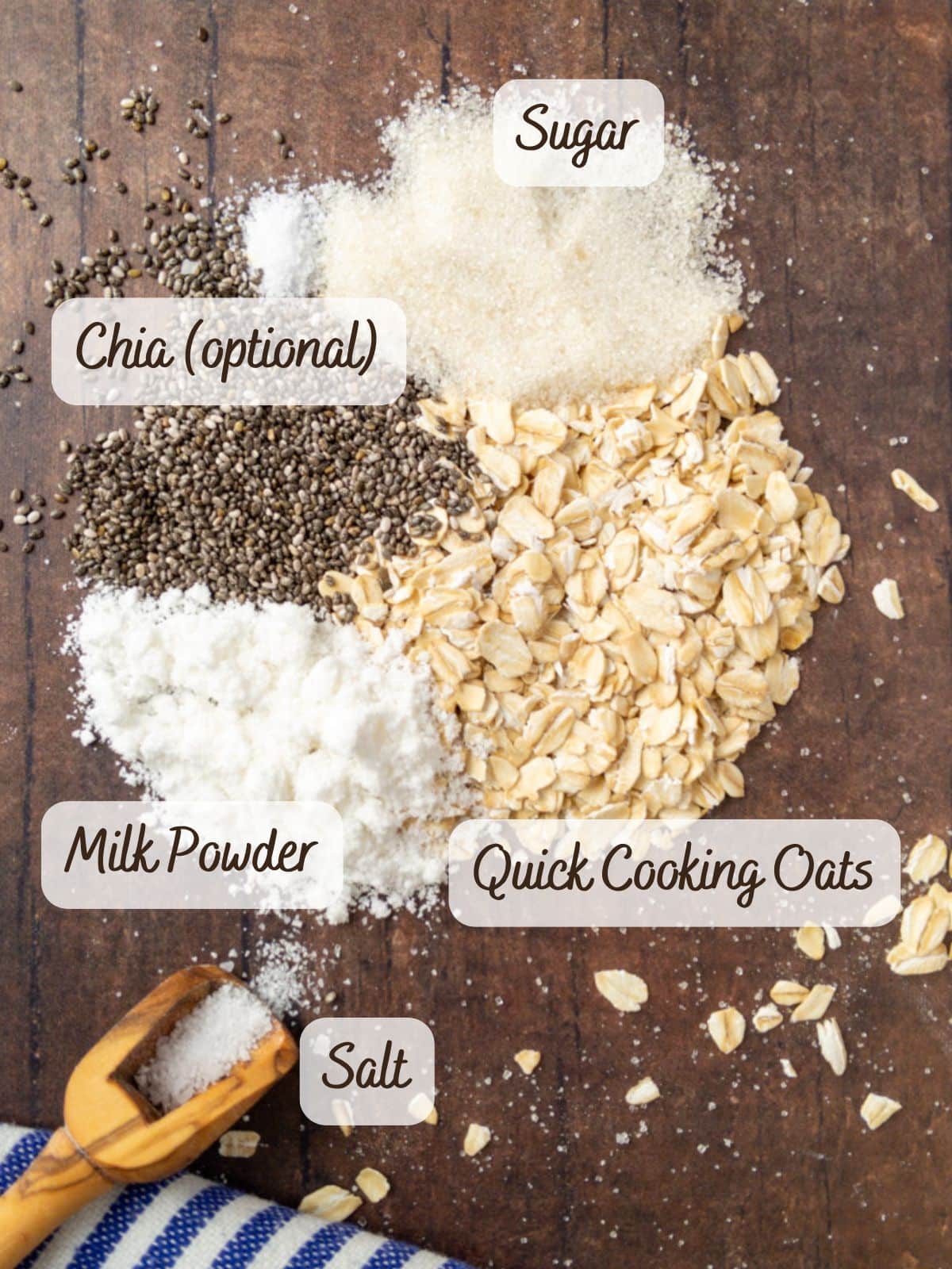 Ingredients for instant overnight oats labeled on a wooden surface: sugar, chia seeds (optional), milk powder, salt, and quick cooking oats. A small scoop of salt and a blue-striped cloth sit nearby, ready to bring this wholesome breakfast to life.