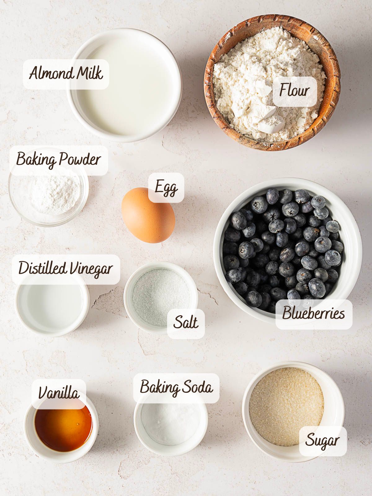 An overhead view of labeled ingredients for baking gluten-free pancakes, including almond milk, flour, baking powder, an egg, blueberries, distilled vinegar, salt, vanilla, baking soda, and sugar on a white surface.