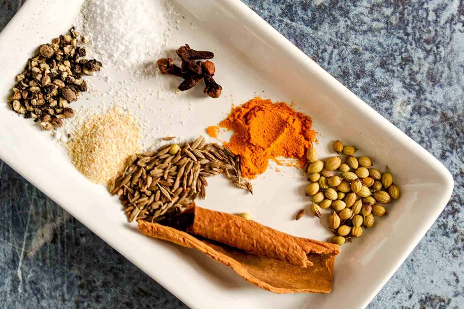A white rectangular plate showcases a medley of spices-pepper, salt, cloves, turmeric powder, garlic powder, cumin seeds, coriander seeds, and a cinnamon stick-all ready to perfectly season succulent chicken wings on a textured surface.