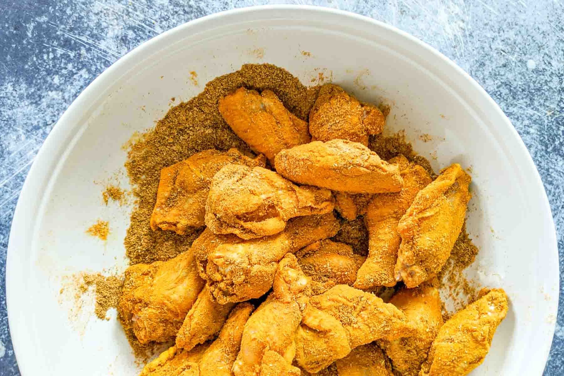 A white bowl holds chicken wing pieces, each coated in a seasoned spice mix, resting on a textured surface.