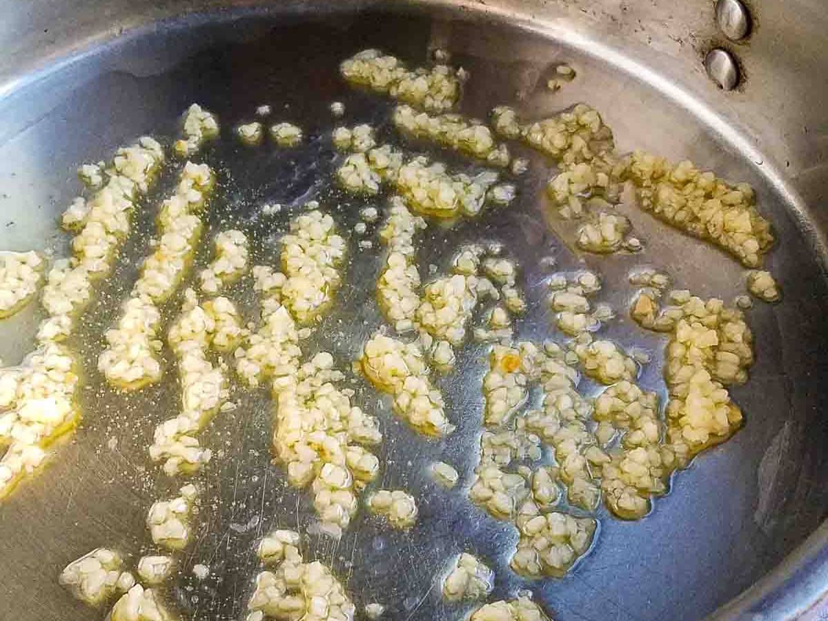Chopped garlic sautéing in oil in a stainless steel pan, perfect for tossing with crisp garlic green beans.
