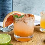 A mezcal paloma in a glass with a chili powder–rimmed edge, garnished with a grapefruit slice and mint. A halved lime and a bottle rest on a wooden surface, while another similar drink is partially visible.