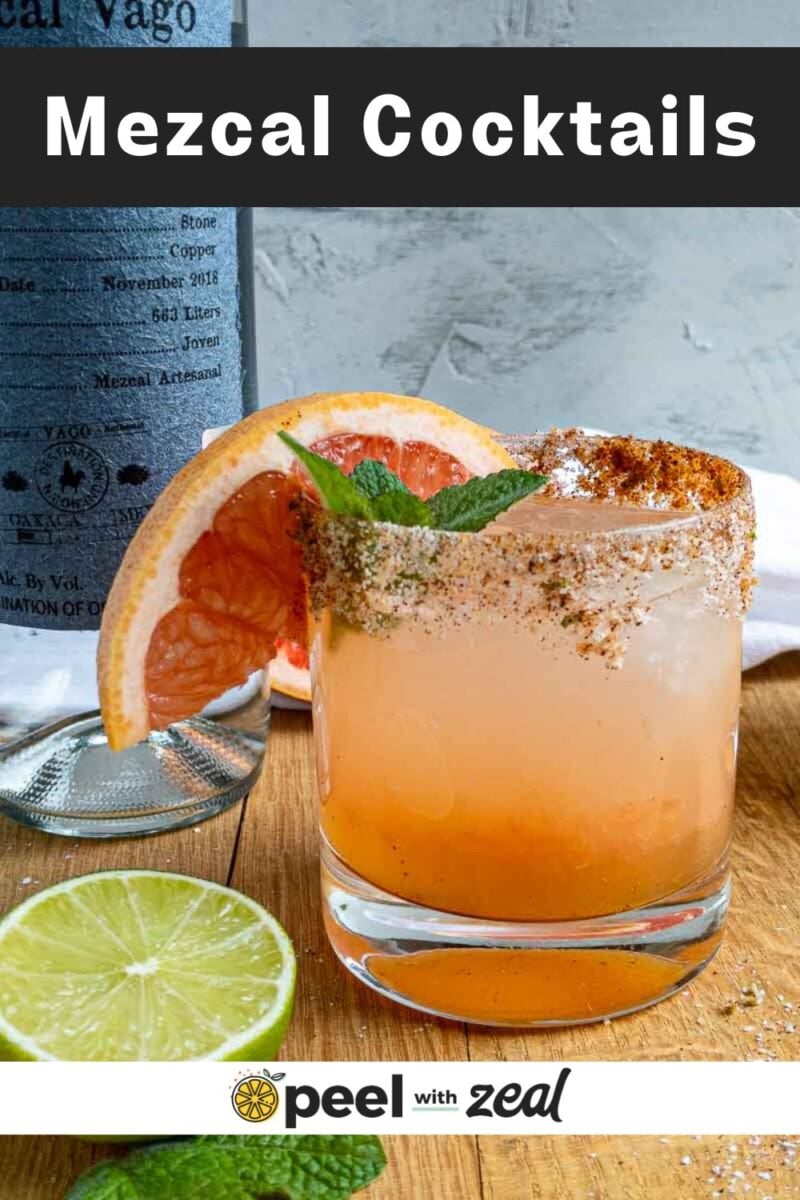 A glass of mezcal paloma cocktail garnished with a grapefruit slice, mint, and a chili-rimmed edge sits on a wooden surface next to sliced lime and grapefruit, with a bottle and printed text in the background.