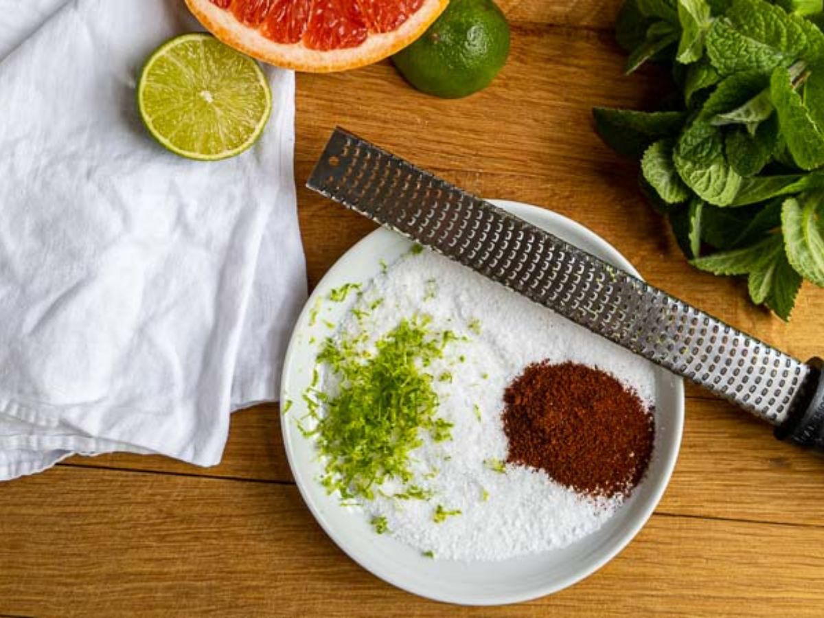 A plate with white granulated salt, chili powder, and lime zest-perfect rimmers for a mezcal paloma-sits on a wooden surface. Nearby are a zester, half a lime, half a grapefruit, fresh mint, and a white cloth.