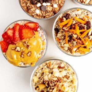 Five bowls of overnight oats topped with various ingredients, including strawberries, peanut butter, chocolate chips, raisins, nuts, and orange zest, arranged on a white surface.