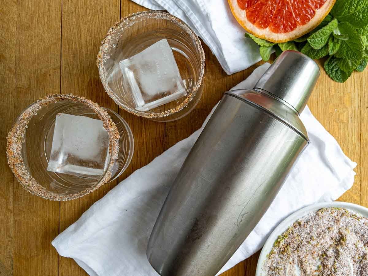 A metal cocktail shaker rests on a white cloth next to two glasses of mezcal paloma with large ice cubes and seasoned rims. A halved grapefruit, mint leaves, and a bowl of seasoning are also visible on a wooden surface.