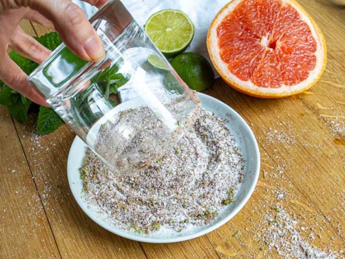 A hand presses a glass into a shallow dish of salt and spices on a wooden surface-perfect prep for a mezcal paloma. Nearby are a halved grapefruit, limes, mint leaves, and a white cloth.