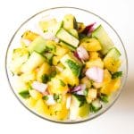 A clear bowl filled with a vibrant pineapple relish salad made of chopped pineapple, cucumber, red onion, and cilantro on a white background.