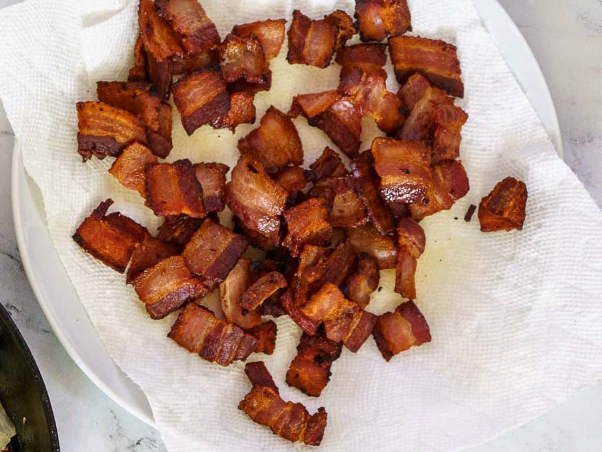 A plate lined with paper towels holds several pieces of cooked, crispy bacon cut into small chunks-perfect for topping your favorite blue cheese potato salad.
