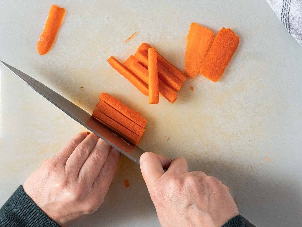 A person slicing a carrot into thin matchstick pieces on a white cutting board with a large knife, preparing fresh ingredients for a vibrant vegan poke bowl.