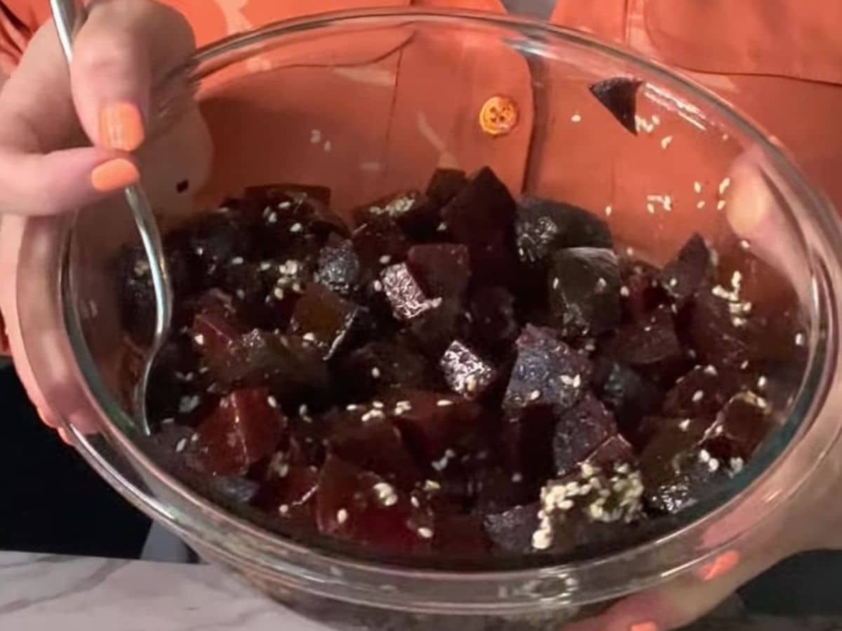 A person holds a glass bowl filled with cubed, dark red food topped with sesame seeds, stirring their vegan poke bowl with a spoon.