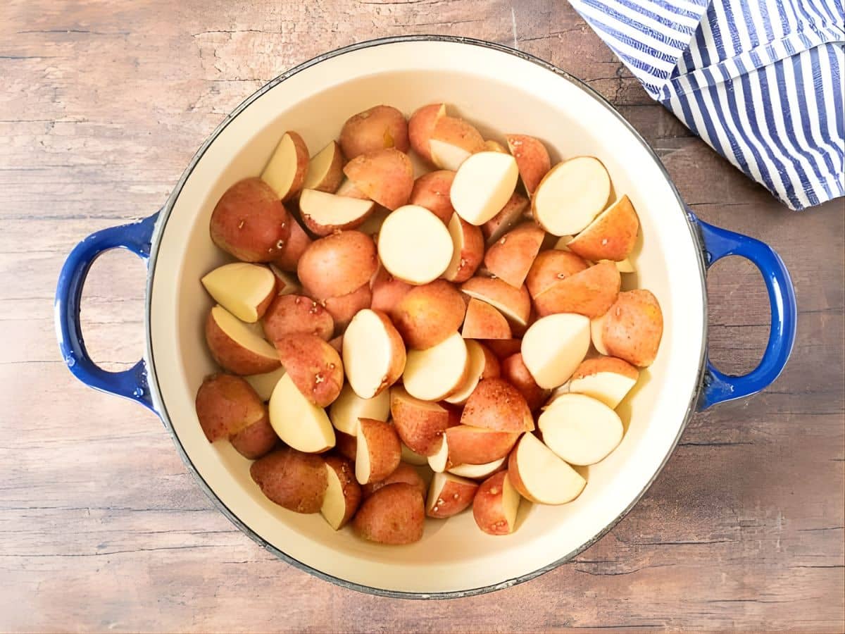 A blue-handled pot filled with halved and quartered red potatoes sits on a wooden surface next to a striped cloth, ready to be transformed into a creamy blue cheese potato salad.