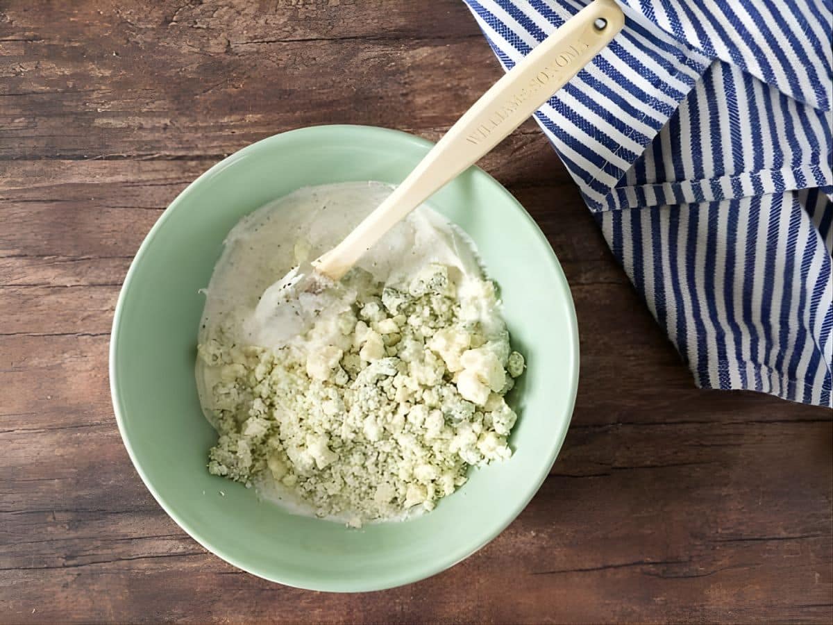 A light green bowl with sour cream and blue cheese being mixed for a blue cheese potato salad, on a wooden surface next to a blue and white striped towel.
