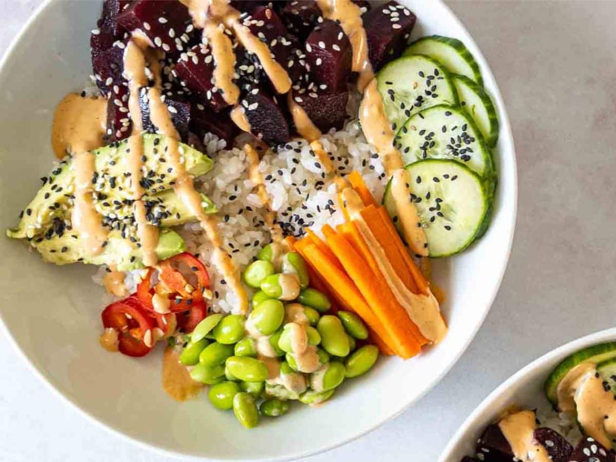 A vegan poke bowl with rice, sliced cucumber, avocado, carrots, edamame, and seaweed, topped with sesame seeds and drizzled sauce.