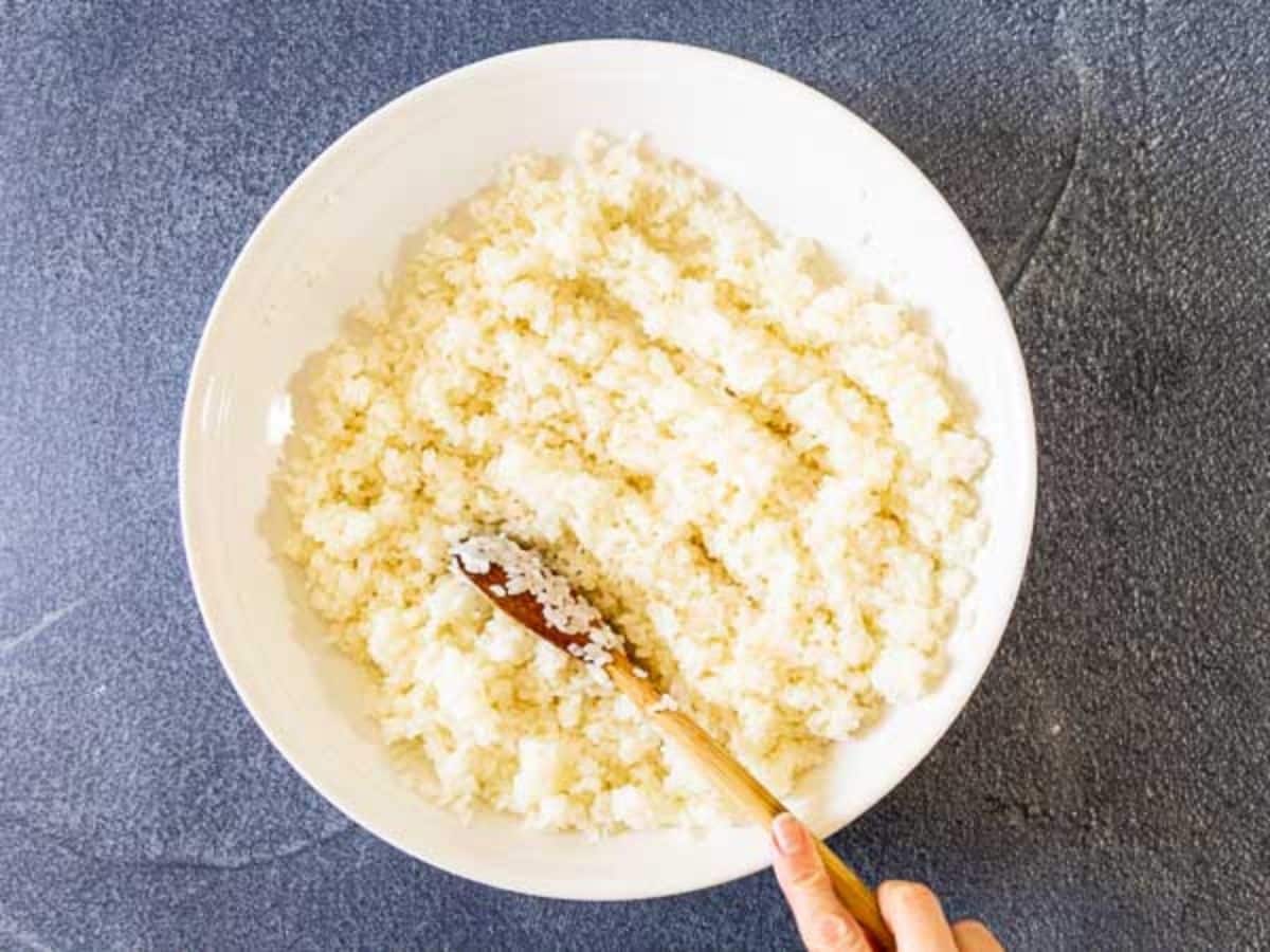 A hand mixes white rice in a large white bowl with a wooden spoon, preparing the base for a delicious vegan poke bowl, set on a dark gray surface.