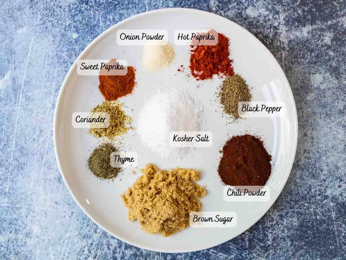 A white plate with labeled piles of spices—perfect for smoked baby back ribs: onion powder, hot paprika, sweet paprika, coriander, black pepper, kosher salt, thyme, chili powder, and brown sugar—rests on a textured blue surface.
