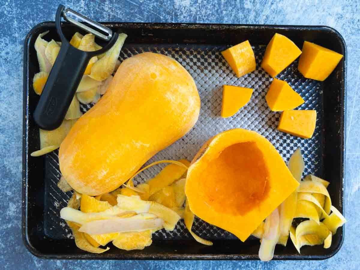 A halved butternut squash, peeled with its seeds removed, sits on a tray with squash cubes, peels, and a black vegetable peeler-perfect for adding to a Vegetarian Thai Green Curry.
