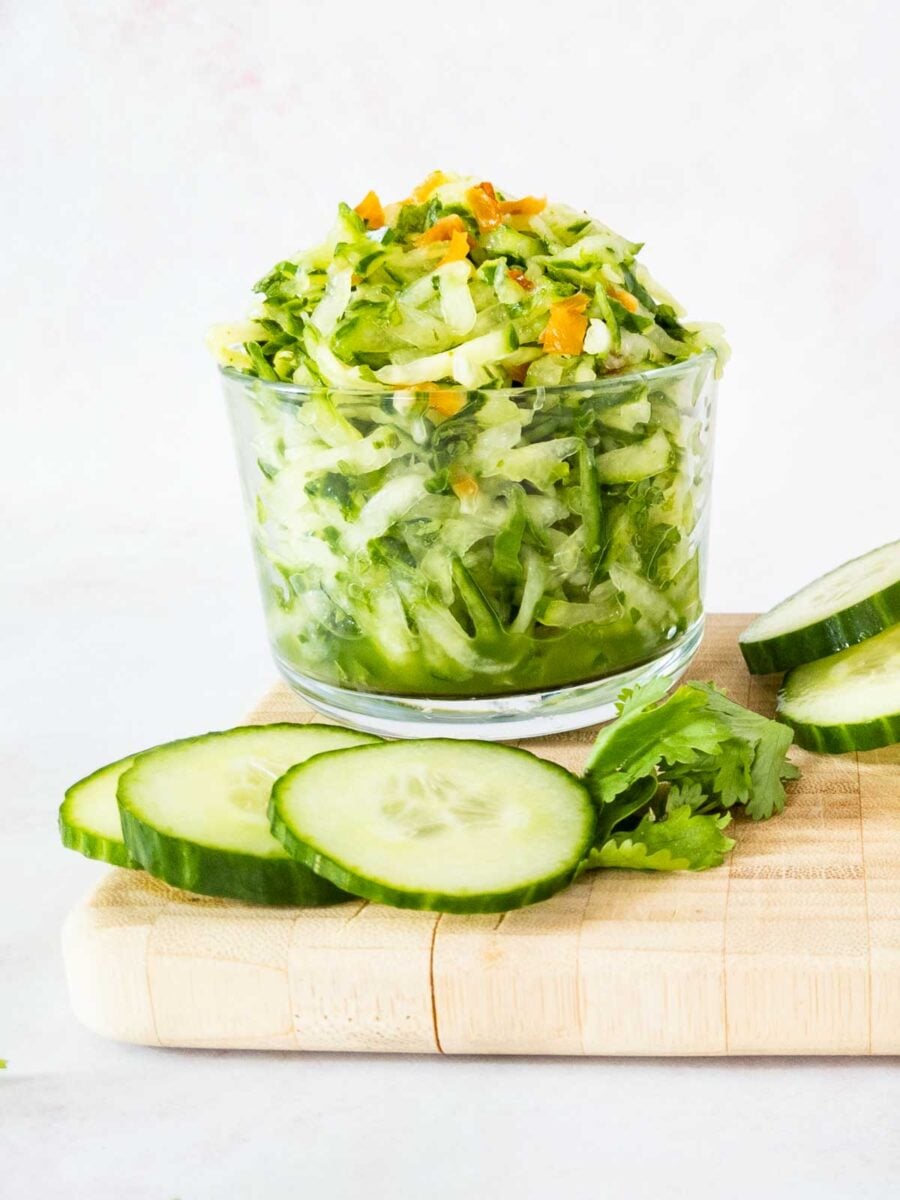 A clear glass filled with shredded cucumber chutney sitting on a wooden cutting board next to fresh cucumber slices and cilantro.