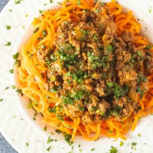 A white plate with orange spaghetti topped with a savory harissa beef sauce and garnished with chopped herbs.