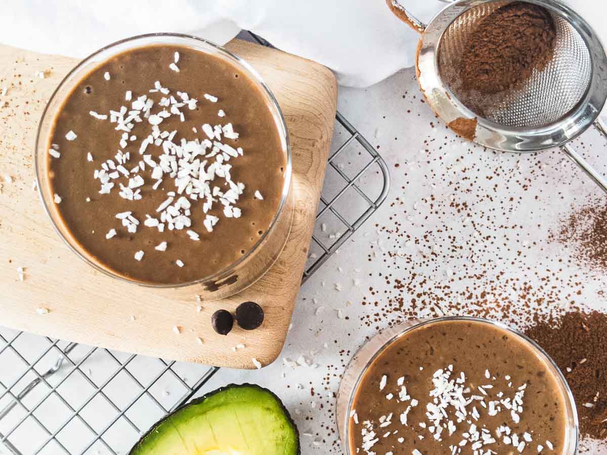 Two chocolate avocado smoothie in glasses with coconut flakes, half of an avocado and cocoa powder.